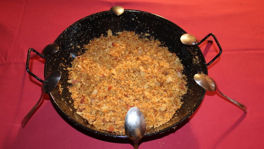 Migas manchegas en sartén con cucharas sobre mesa roja.